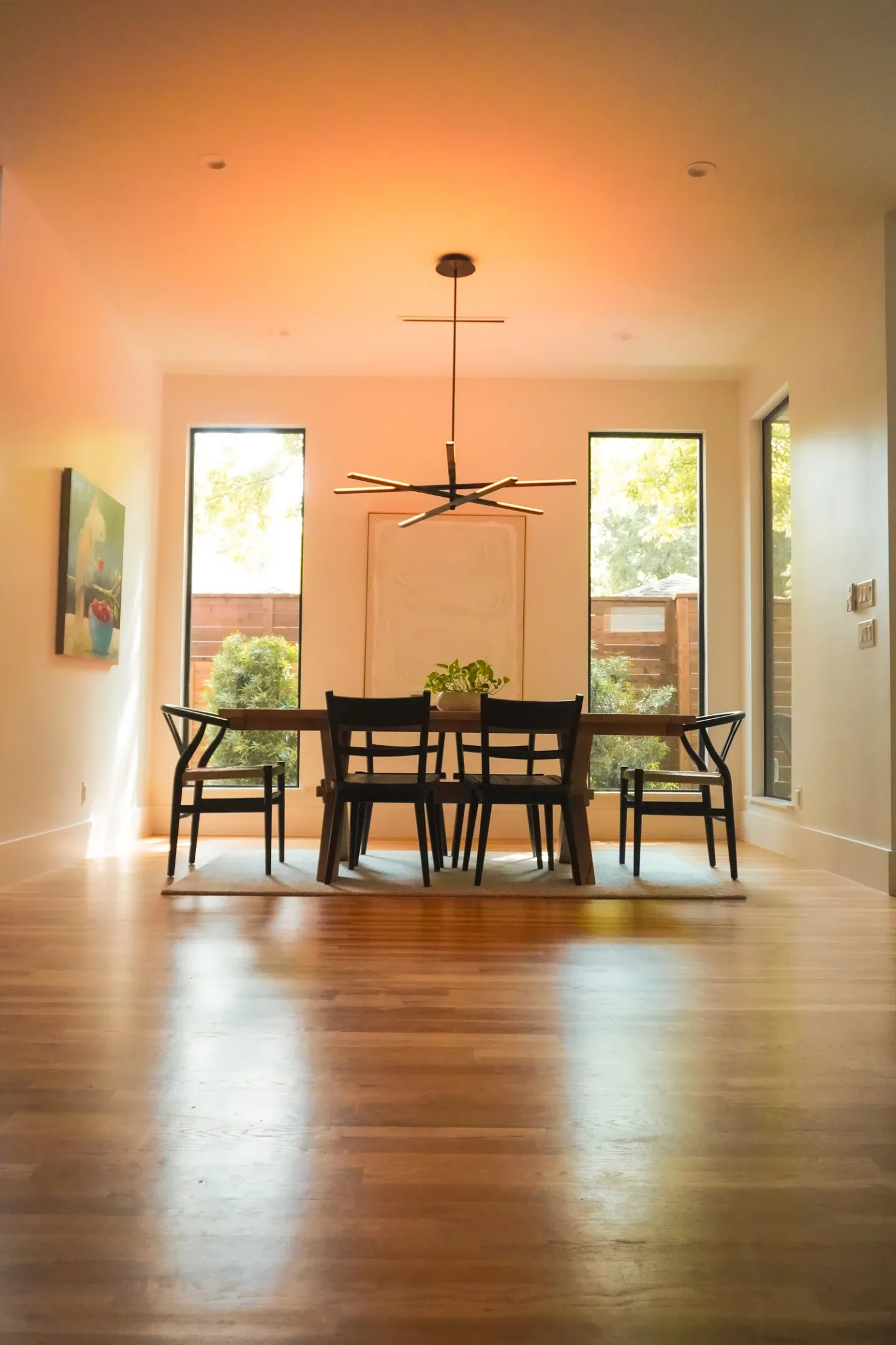 Dining room with a wooden table and six black chairs, a modern black chandelier hanging from the ceiling, two large windows with outdoor views, a plant on the table, and a colorful painting on the wall.
